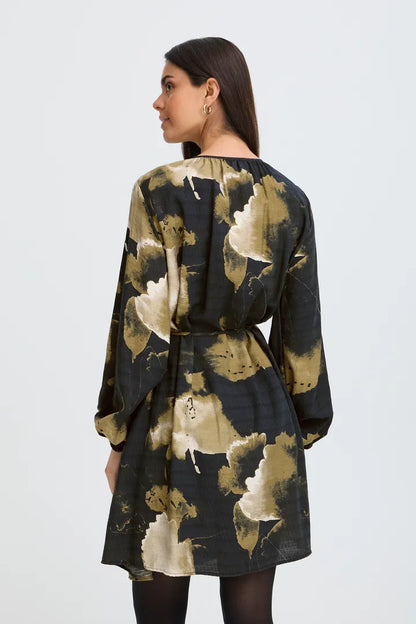 A woman shown from the back wearing a long-sleeve tunic dress with a black base and a large abstract floral print in beige and olive green, paired with black tights against a light gray studio background.
