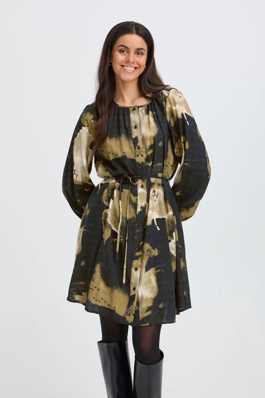 A woman modeling a long-sleeve mini dress with a black and olive green abstract tie-dye pattern, featuring a drawstring waist and front pockets, paired with black tights and black boots against a light gray background.
