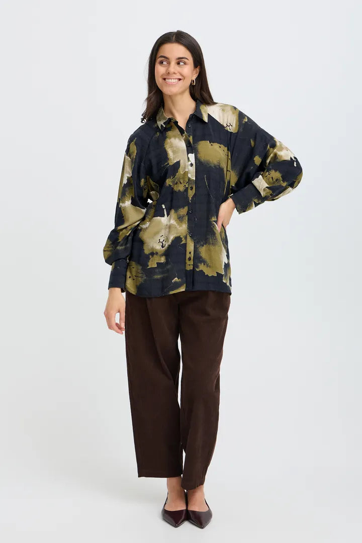 A woman modeling a long-sleeve button-up shirt with a navy and olive green abstract pattern, paired with dark brown trousers and black pointed-toe flats, against a light gray studio background.