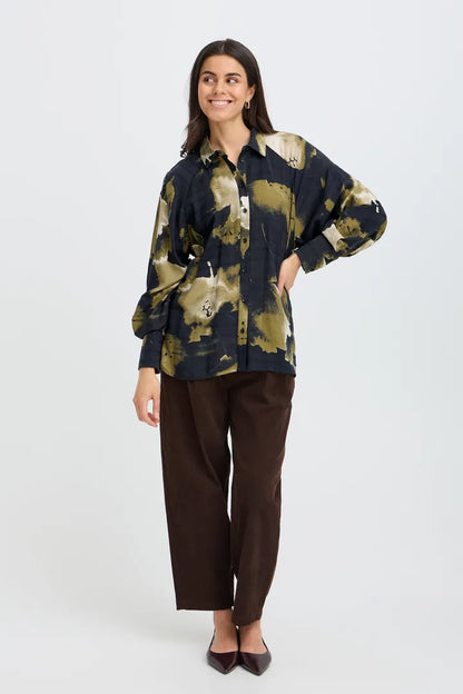 A woman modeling a long-sleeve button-up shirt with a navy and olive green abstract pattern, paired with dark brown trousers and black pointed-toe flats, against a light gray studio background.