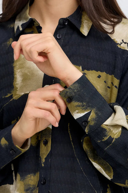 Close-up of a person wearing a long-sleeve button-up shirt with a dark navy base and large abstract olive-green and beige brushstroke pattern, adjusting the cuff.
