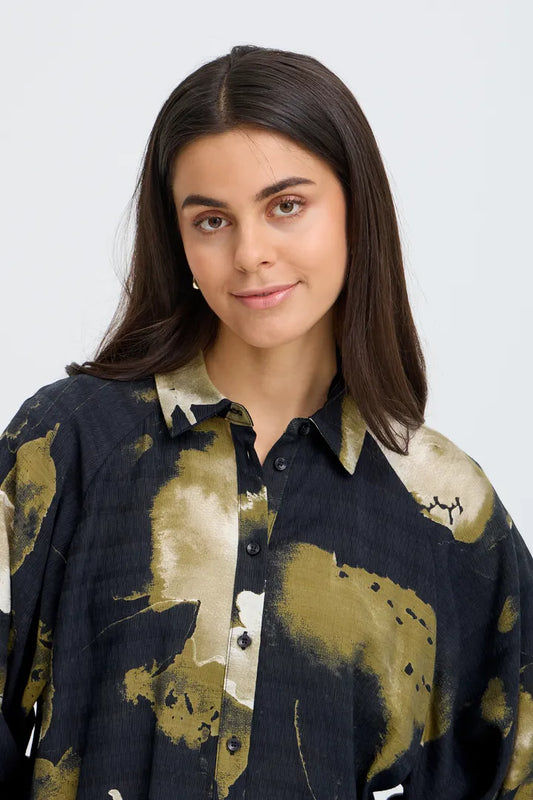 A woman with dark brown hair wearing a dark navy button-up shirt with a gold and beige abstract paint-splatter pattern, photographed against a light gray background.