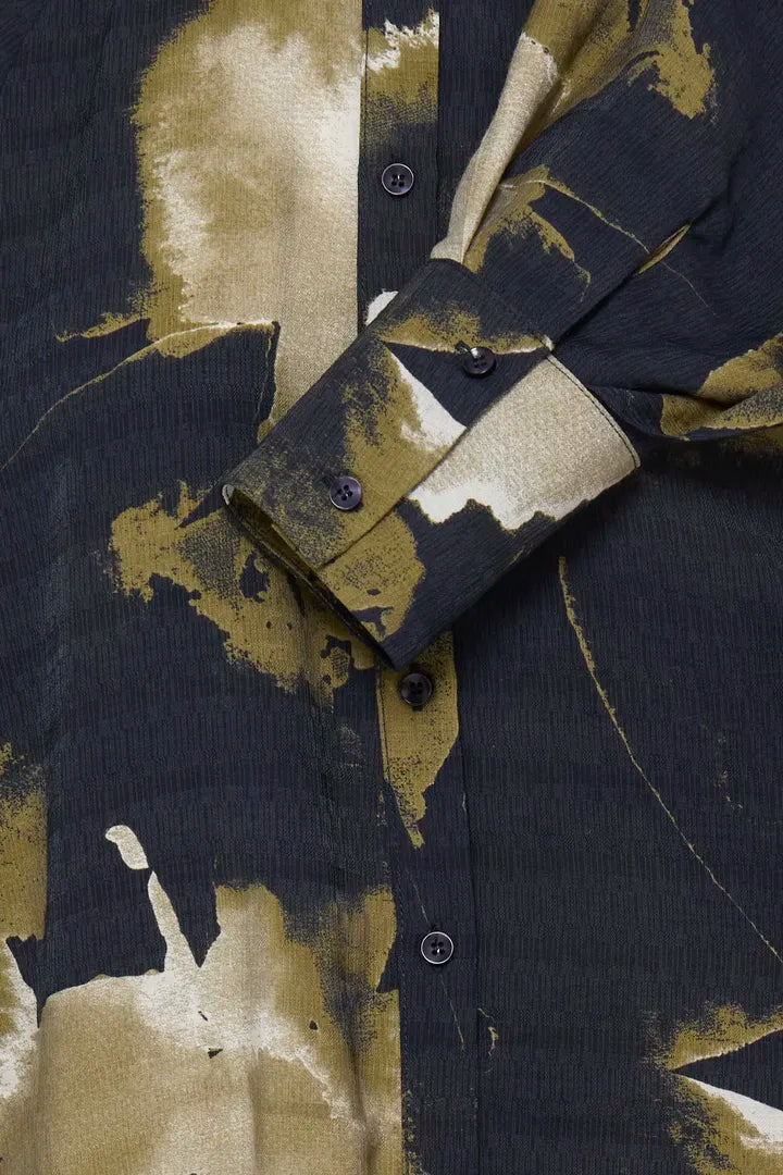 Close-up of a button-front shirt with a dark charcoal base and large abstract brushstroke pattern in beige and olive-gold tones. The cuff is folded over, showing dark buttons and the fabric texture.