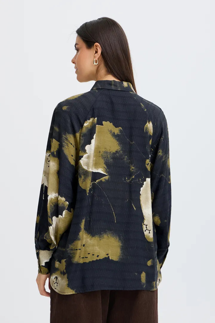 A woman shown from the back wearing a dark navy long-sleeve shirt with an abstract olive green and cream paint-splatter pattern, paired with brown pants, against a light gray studio background.