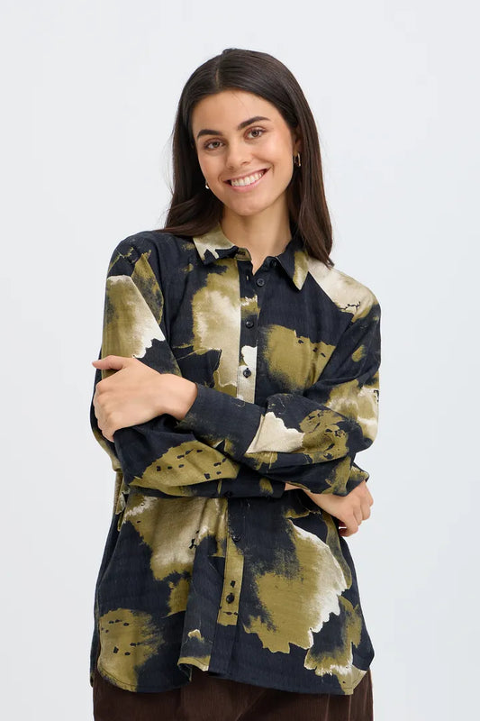 A woman modeling a long-sleeve button-up shirt with an abstract watercolor-style print in olive green, beige, and dark navy blue, set against a plain light gray background.